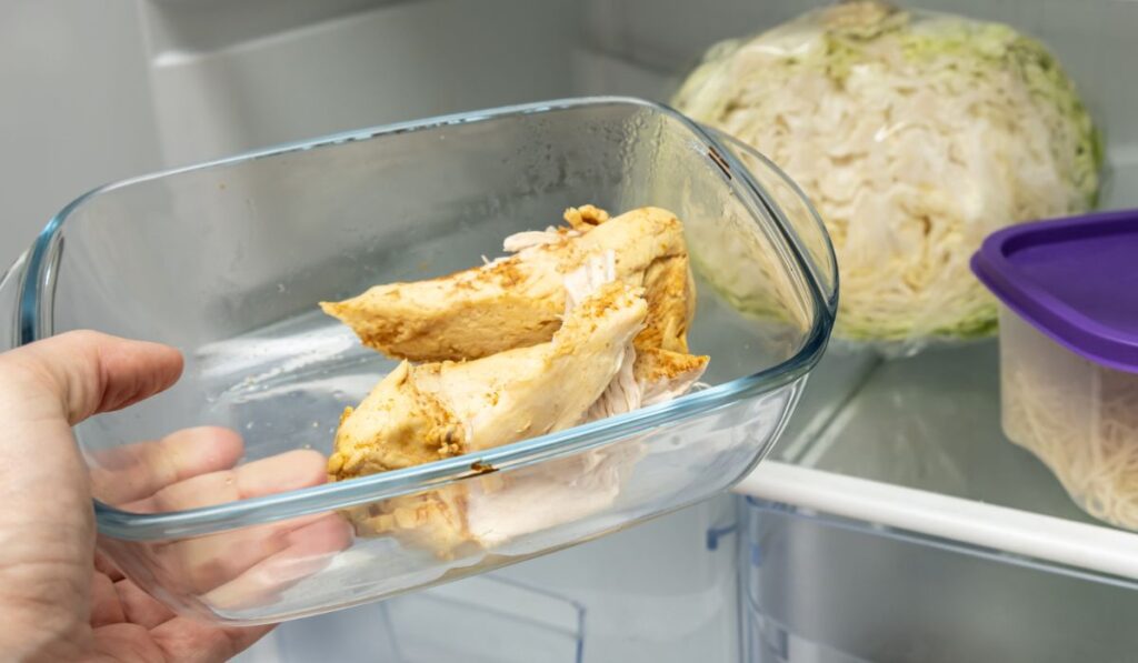 Hand of a dieting person holding taking cooked chicken breasts in a glass container from a fridge